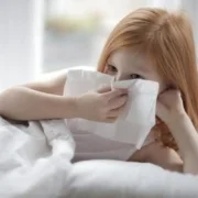young girl with red hair laying in bed with white blankets and blowing her nose