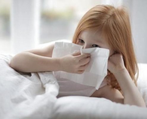young girl with red hair laying in bed with white blankets and blowing her nose
